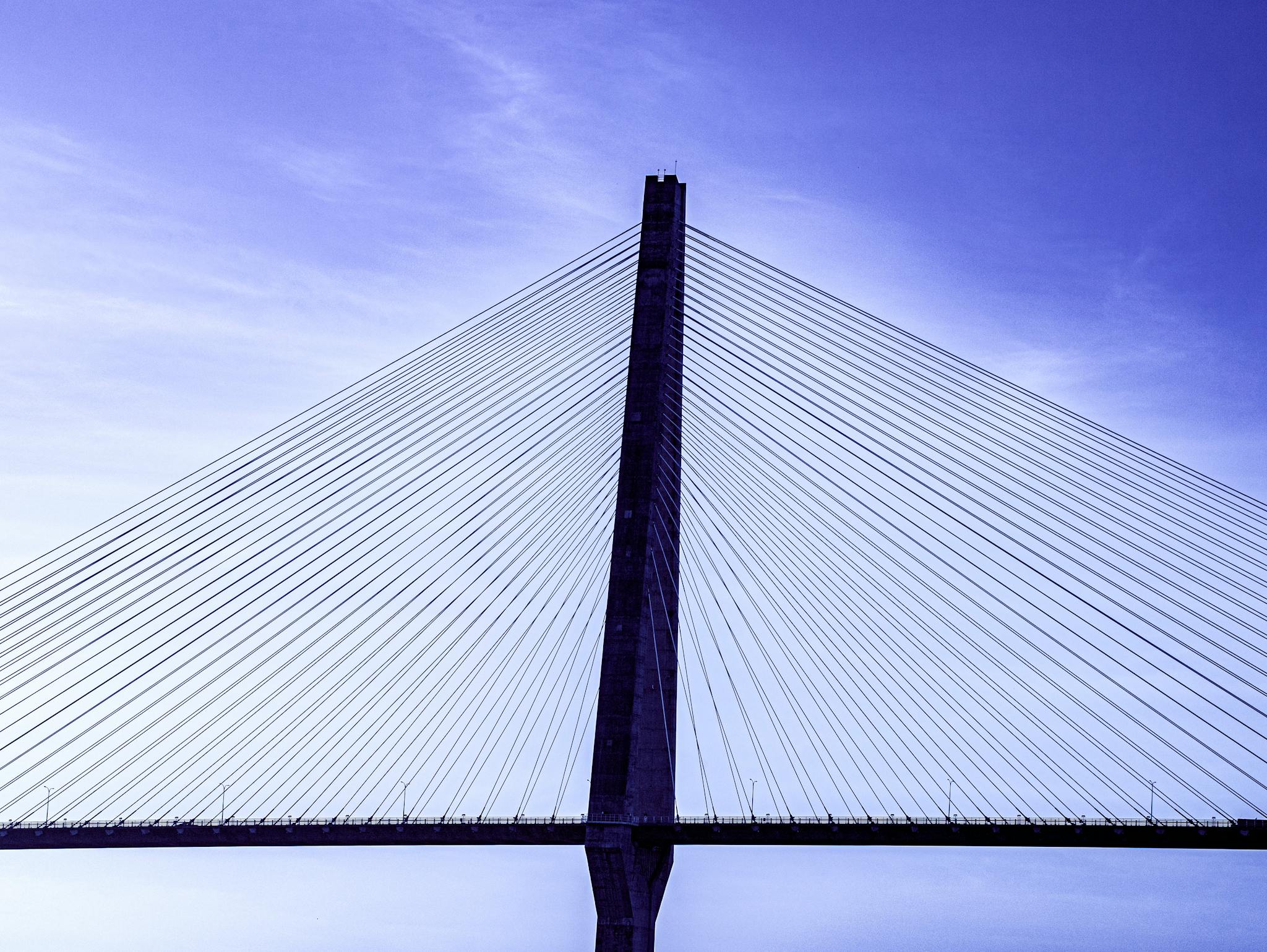 A modern cable-stayed bridge viewed from below with a clear blue sky backdrop.