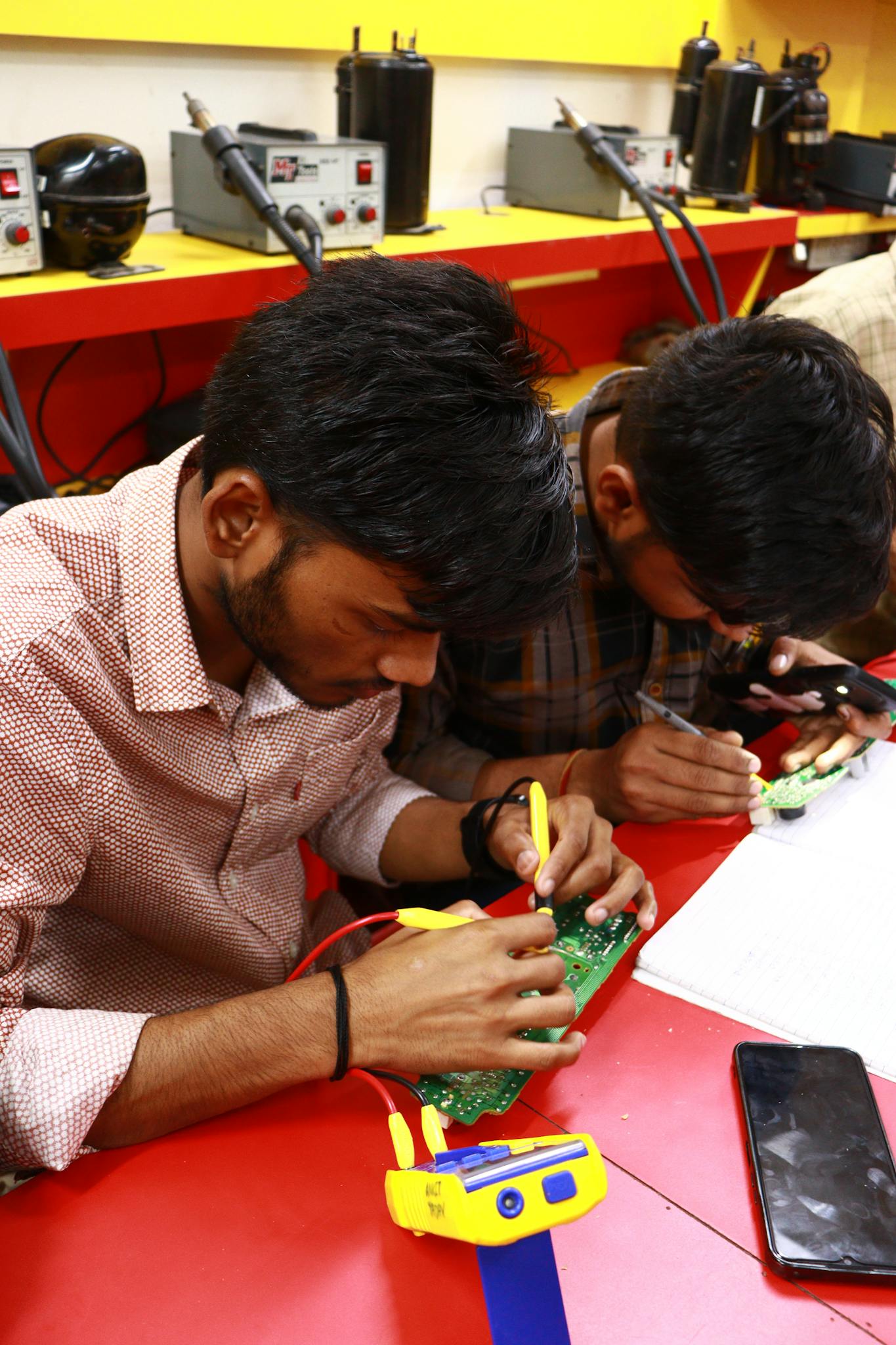 Young men learning PCB repair technique in electronic workshop.